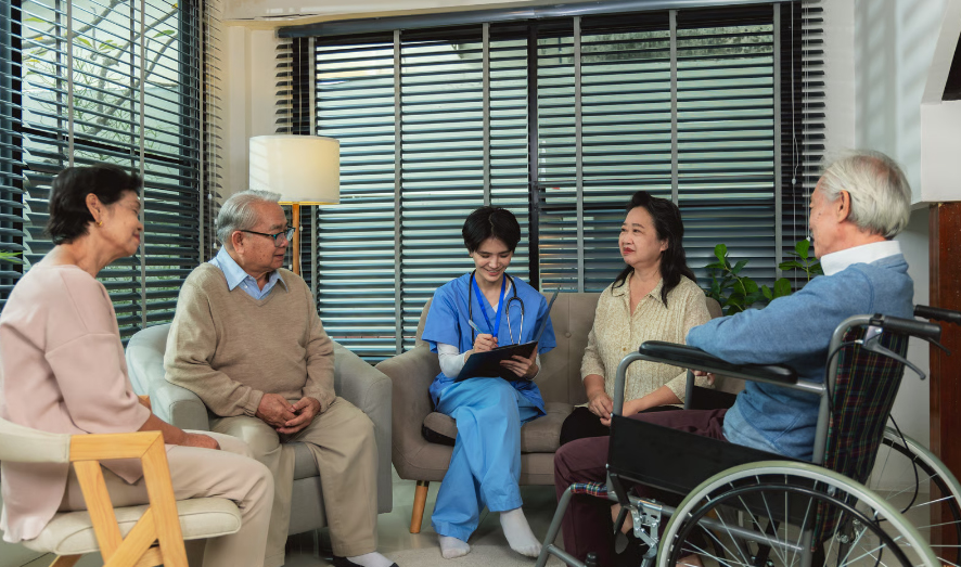 An in-home nurse in blue scrubs takes notes while sitting on a couch, surrounded by two senior couples, one elderly man is in a wheelchair.