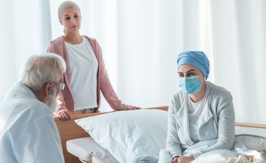 A senior male physician in a white coat speaks with a young female patient wearing a surgical mask and headscarf, while a concerned caregiver looks on in a clinical setting.