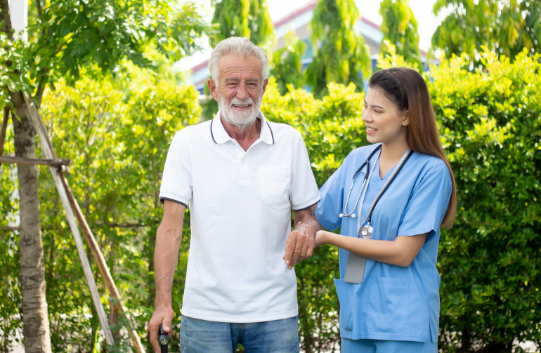 A smiling senior man with a walking cane supported by a young nurse in blue scrubs in a sunny outdoor garden.