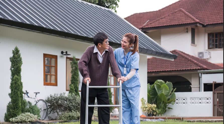 A hospice nurse in blue scrubs smiling and assisting an elderly man using a walker outside his home.