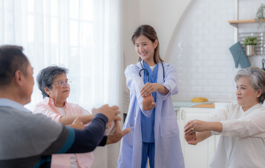 A young female nurse in a white coat and blue scrubs leading three senior citizens in a gentle arm-stretching exercise.