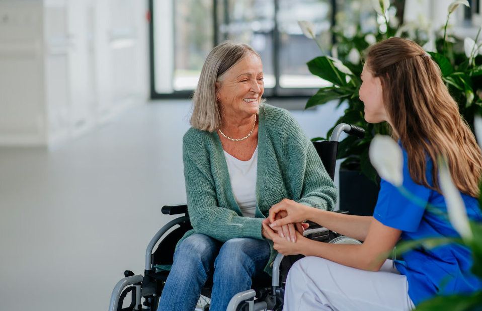 A happy senior woman sitting in a wheelchair holding hands and talking with a caring female home health aide dressed in blue scrubs in a brightly lit care facility.