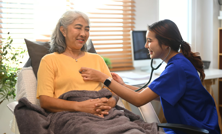 A female home health nurse in blue scrubs uses a stethoscope to check the heartbeat of an elderly woman resting comfortably in a chair.