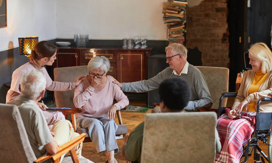An elderly woman cries during a senior support group therapy session while a counselor and another senior man comfort her with hands on her shoulders.