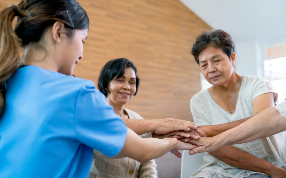 A female hospice nurse in blue scrubs stacking hands with two elderly women, demonstrating the compassionate, team-based support provided to families managing advanced dementia care at home.
