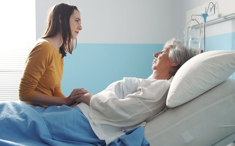 A young woman sits by the bedside of an elderly female hospice patient, gently holding her hand to offer comfort and support.