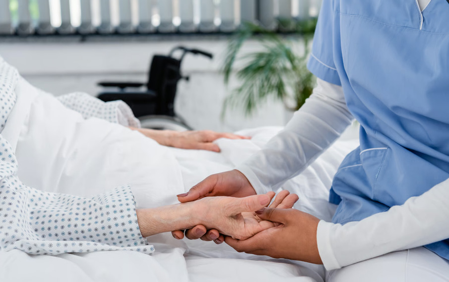 Close-up of a hospice nurse wearing blue scrubs gently holding the hand of a patient resting in a medical bed, with a wheelchair blurred in the background.