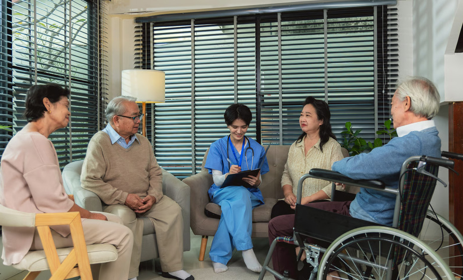 A female hospice nurse in blue scrubs reviewing notes on a clipboard during a face-to-face family consultation with an elderly man in a wheelchair and his loved ones, demonstrating a routine hospice recertification check-in at home.