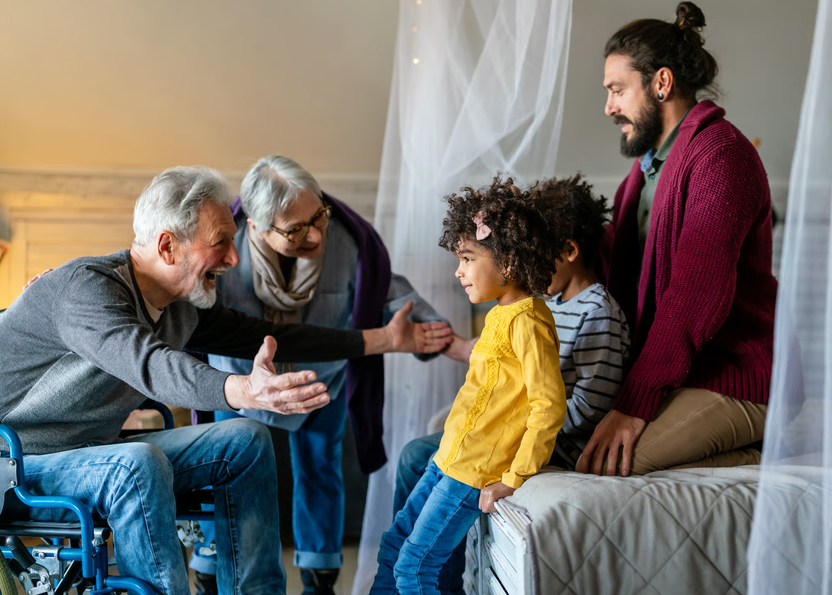 A multi-generational family happily gathering in a bedroom around an elderly man sitting in a wheelchair.