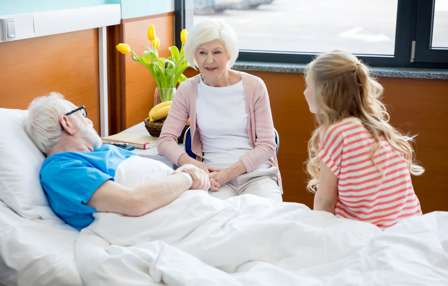 An elderly woman and a young girl visiting a senior man resting in a hospice care bed.