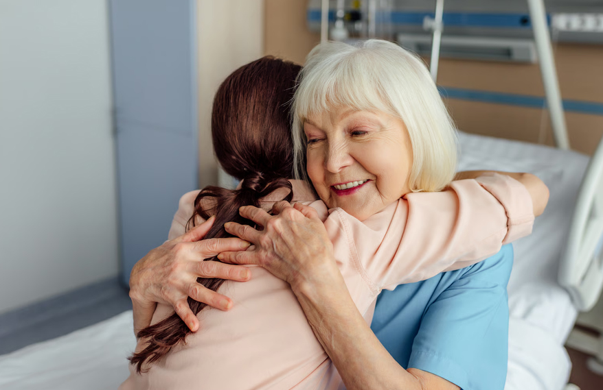 A family caregiver warmly embracing an smiling senior woman sitting on a medical bed, representing relief and emotional support through respite care.