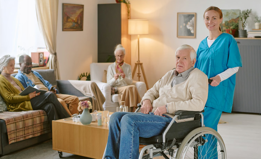 A professional hospice nurse in blue scrubs standing behind a senior man seated in a wheelchair inside a comfortable living area.