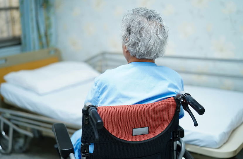 Rear view of an elderly individual with grey hair sitting in a wheelchair next to a medical bed.
